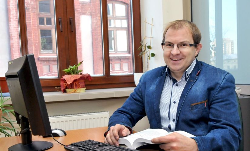 Zdjęcie portretowe: prof. Grzegorz Szymański w niebieskiej marynarce siedzi przy biurku i komputerze.