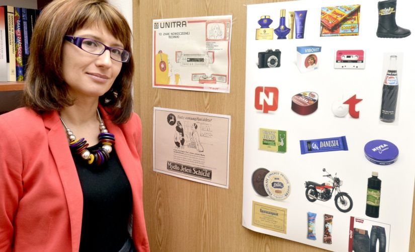 Portrait photo: smiling Assoc. Prof. Eng. Magdalena Grębosz-Krawczyk, TUL Prof. wearing a red jacket, is standing next to a cork board with colourful pictures.