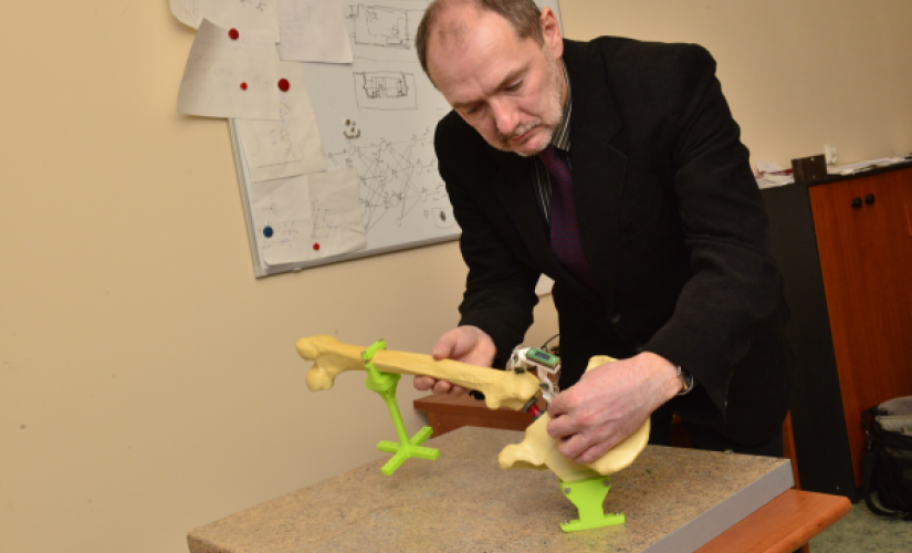Zdjęcie portretowe: prof. Leszek Podsędkowski stoi przy biurku i składa model z żółtych i zielonych elementów. w tle tablica z dokumentami.