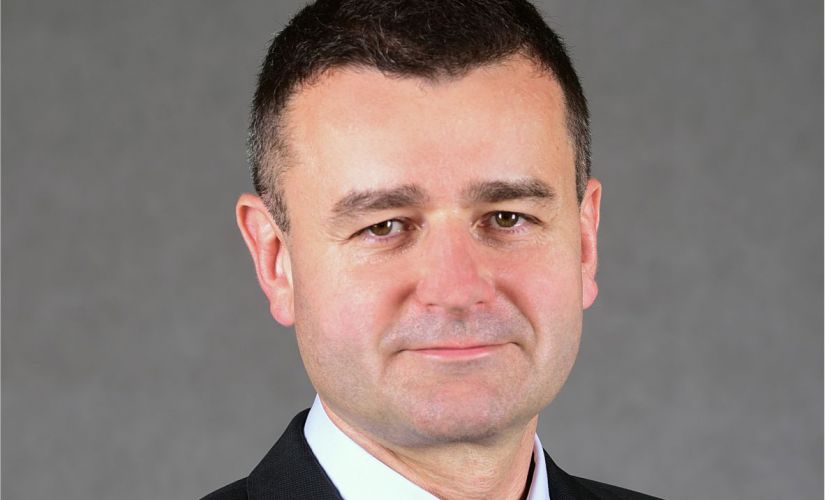 Zdjęcie portretowe: prof. Andrzej Romanowski w ciemnym garniturze, białej koszuli i niebieskim krawacie na szarym tle.