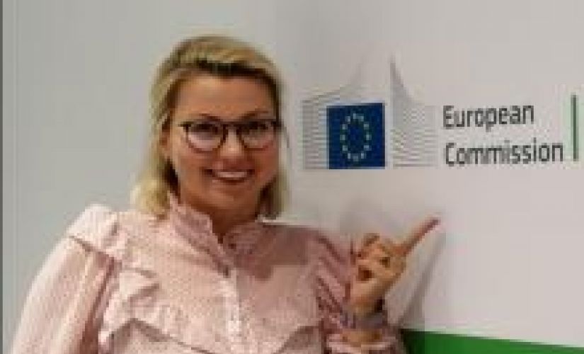 Zdjęcie portretowe: uśmiechnięta Kaja Kantorska na tle jasnej ściany z logiem European Comission, które wskazuje palcem wskazującym.