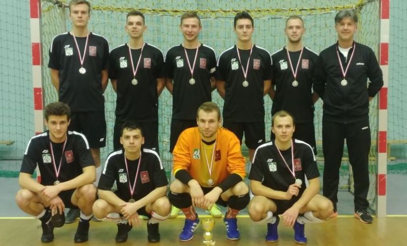 9-osobowa drużyna futsalu PŁ z trenerem S. Ścieszko stoją na tle bramki w dwóch rzędach.