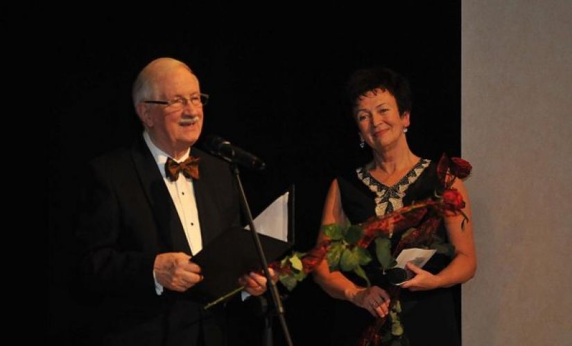 Prof. Jan Krysiński w smokingu i Grażyna Sikorska w czarnej sukni na scenie stoją przy mikrofonie na czarnym tle.