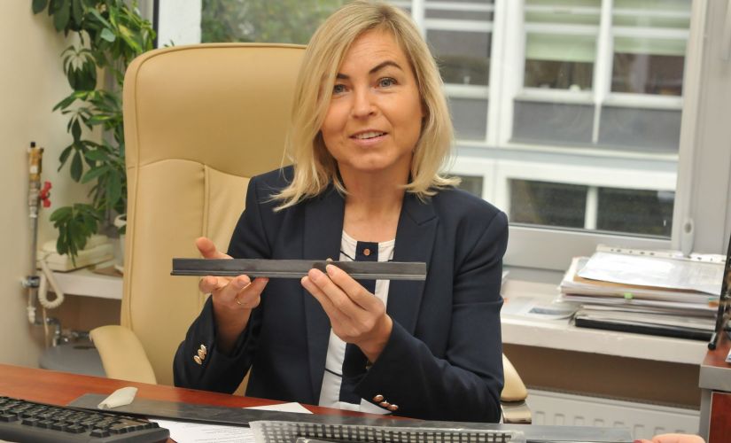 Zdjęcie portretowe. Uśmiechnięta prof. Renata Kotynia siedzi przy biurku w swoim gabinecie. W dłoniach trzyma metalowy element konstrukcji.
