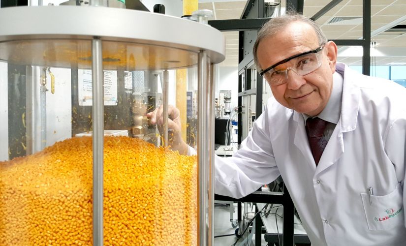 Zdjęcie portretowe: Prof. Andrzej Górak w białym fartuchu i okularach ochronnych stoi przy przeźroczystym pojemniku wypełnionym żółtymi granulkami.