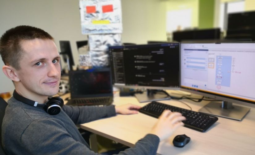 Łukasz Wiśniewski, absolwent PŁ, siedzi przy biurku na którym stoją 2 czarne monitory i klawiatura.