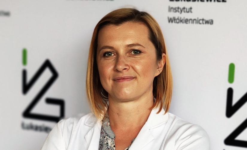 Zdjęcie portretowe: Magdalena Olak-Kucharczyk na tle ścianki z logo Sieć Badawcza Łukasiewicz.