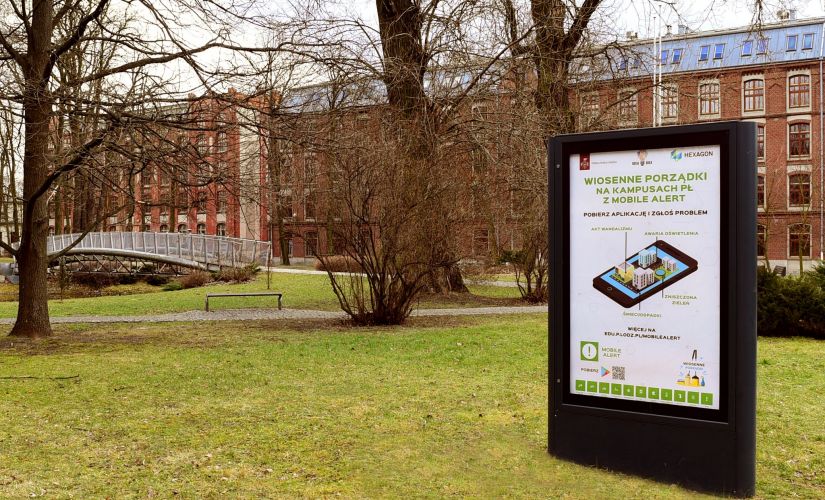 Kampus B Politechniki Łódzkiej: na pierwszym planie citylight z plakatem reklamowym. W tle park z niewielką ilością zieleni oraz Budynek Trzech Wydziałów.