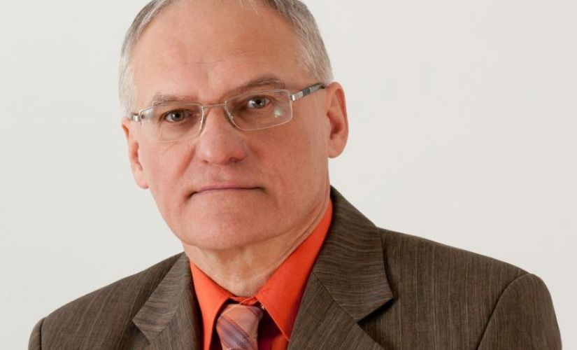 Zdjęcie portretowe: prof. Jan Awrejcewicz w szarym garniturze, łososiowej koszuli i krawacie na jasnej ścianie.