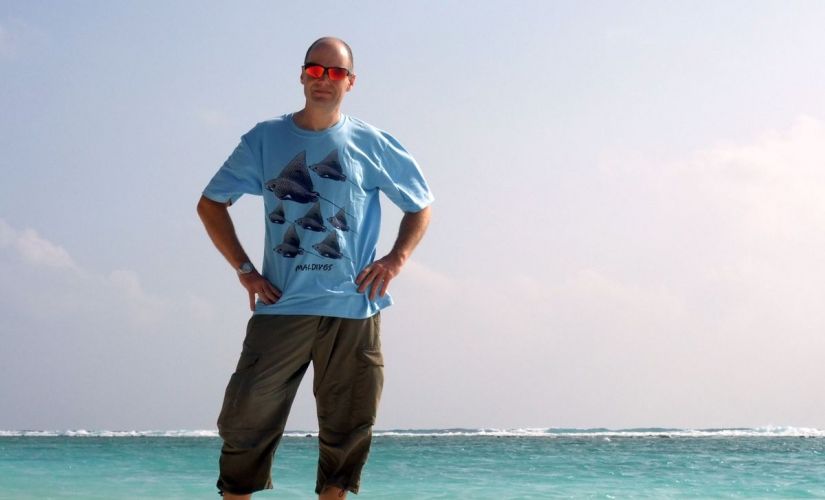 prof. K. Olejnik w przeciwsłonecznych okularach, niebieskim t-shircie i brązowych spodniach stoi na skraju morza.