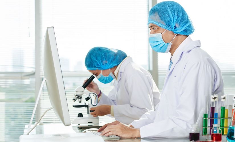 Laboratorium chemiczne. Dwoje badaczy w białych fartuchach i niebieskich czepkach i maseczkach w laboratorium stoją przy mikroskopie i ekranie.