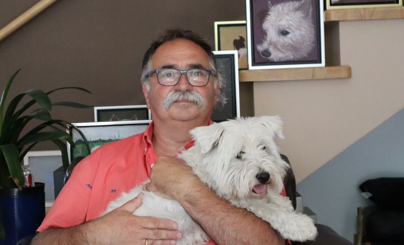 Prof. Tomasz Kapitaniak w łososiowej polówce siedzi w fotelu i trzyma na rękach białego psa. W tle ściana ze zdjęciami tego samego psa.