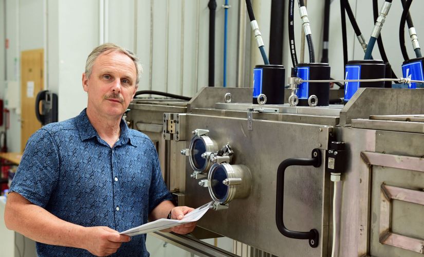 Dr inż. Adam Rylski w niebieskiej koszuli z krótkim rękawie w laboratorium, przy srebrnej maszynie.