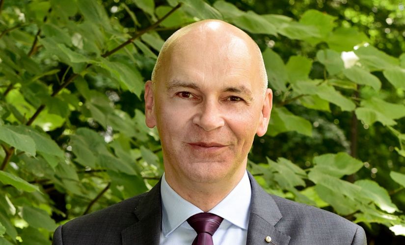 dr hab. inż. Witold Pawłowski, prof. PŁ w szarym garniturze, białej koszuli i bordowym krawacie na tle zieleni.