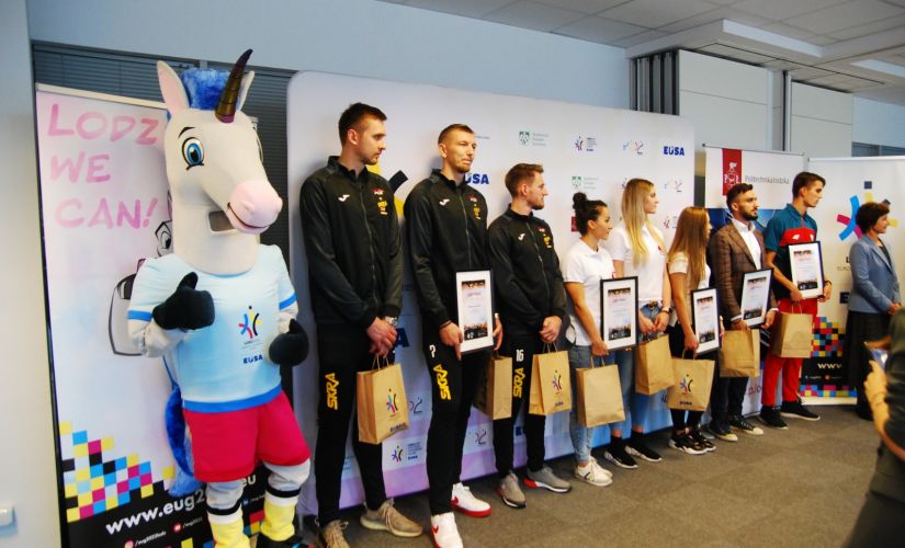 Od lewej strony, w jednym rzędzie stoją: maskotka EUG2022 - kolorowy jednorożec i ośmiu ambasadorów wydarzenia w strojach sportowych.