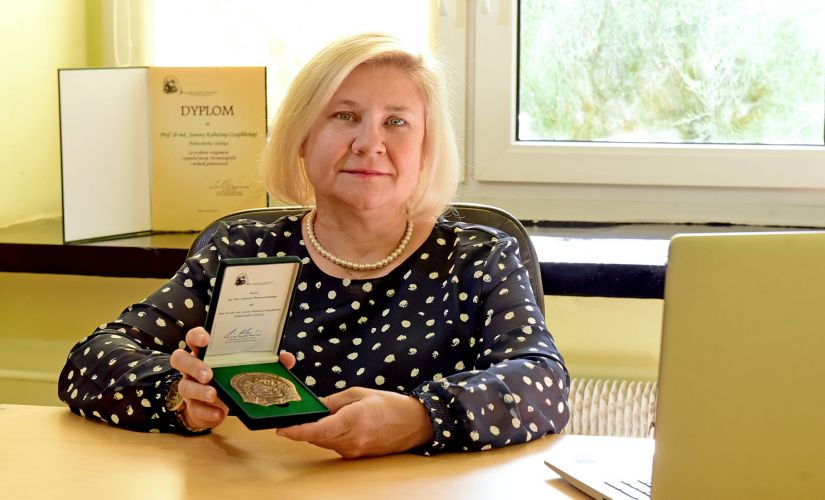 Zdjęcie portretowe: prof. Joanna Czaplińska-Kałużna siedzi przy biurku i prezentuje przyznanym jej Medalem im. Prof. Andrzeja Waksmundzkiego.