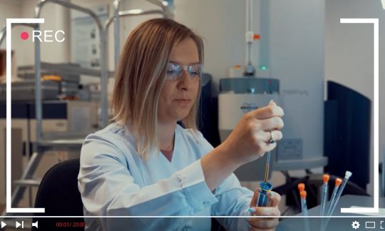 Kadr z filmu: laborantka w białym fartuchu przy stanowisku badawczym.