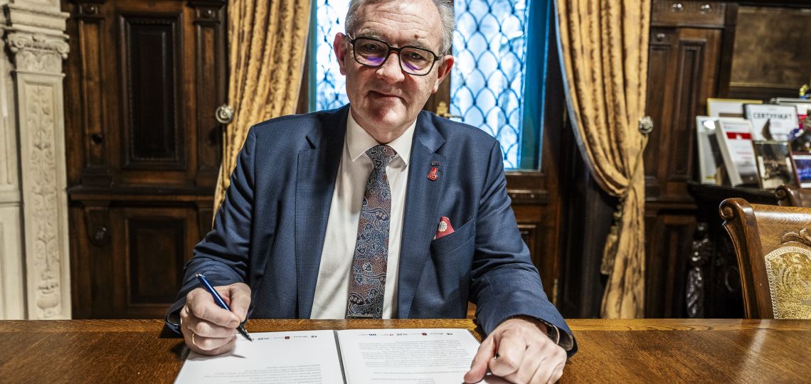 patrzący w obiektyw mężczyzna w okularach i garniturze siedzi przy stole i podpisuje dokumenty piórem