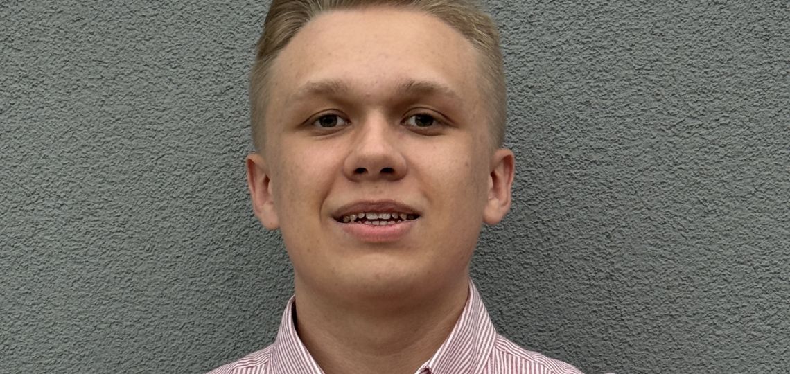 portret młodego mężczyzny o blond włosach, opierającego się o szarą ścianę, ma na sobie koszulę w biało-r&oacute;żowe pionowe paseczki
