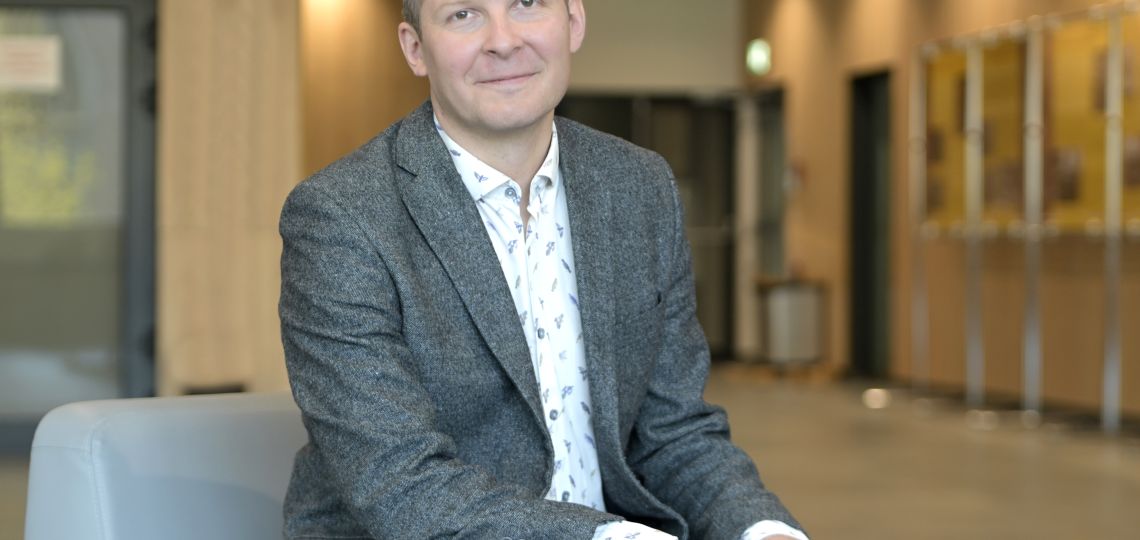 mężczyzna w średnim wieku uśmiecha się na zdjęciu, siedząc z dłońmi na kolanach opartymi