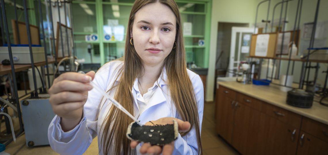 studentka w białym fartuchu trzyma pipetę oraz kawałek ciemnego materiału, na którym widać skroploną wodę, w tle zyblurowane laboratorium