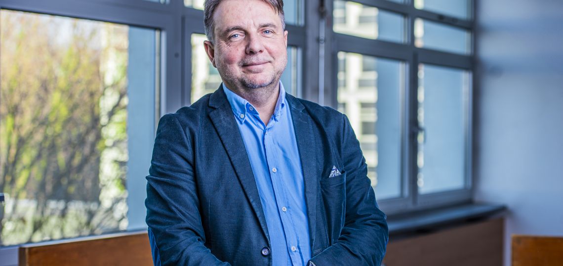 Mężczyzna w granatowej marynarce i błękitnej koszuli stoi na tle ściany z oknami