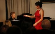 Śpiewaczka w czerwonej sukni stoi i śpiewa przy fortepianie przy którym siedzi akompaniatorka.
