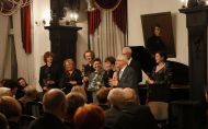 Elegancko ubranych sześciu wykonawców, prowadząca i prof. Jan Krysiński stoją przed widownią po zakończeniu koncertu z cyklu Muzyka na Politechnice. W tle nastrojowa sala, czarny fortepian, a na ścianie portret F. Chopina.