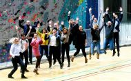 About 30 members of the EUG Lodz 2022 Organising Committee jump up. In the background a climbing wall of the Sports Bay 