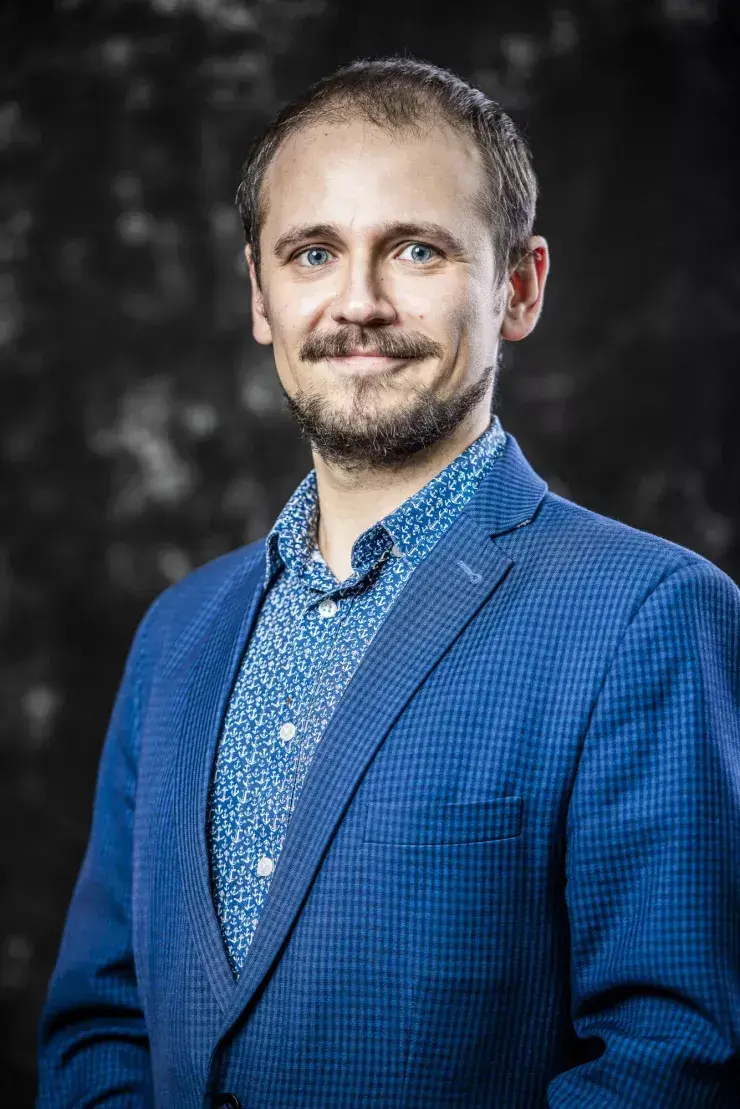 portret mężczyzny w średnim wieku, w granatowej marynarce i koszuli, uśmiecha się, ma brodę, ciemne tło