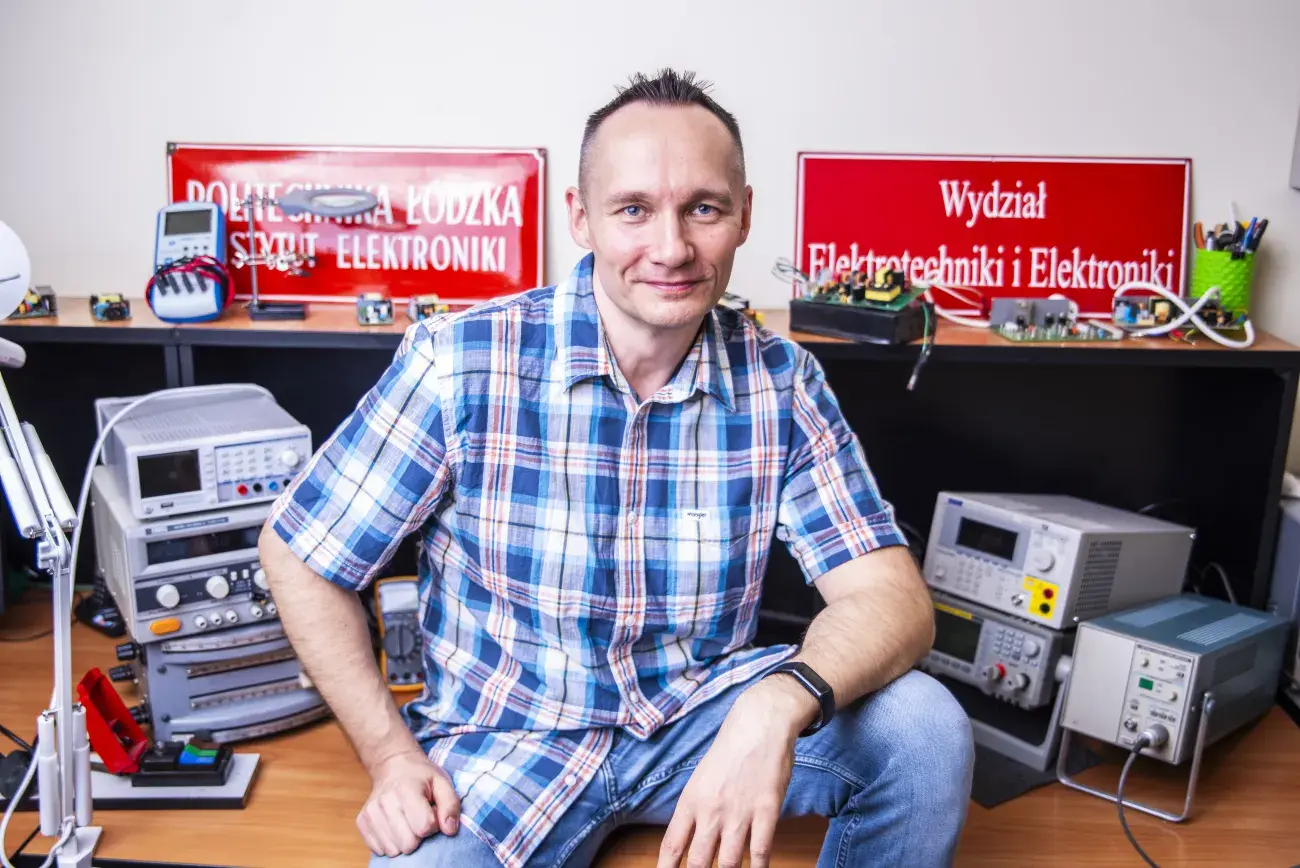 uśmiechnięty mężczyzna w koszuli w kratkę opiera się o biurko, na którym stoją urządzenia elektroniczne, w tle ściana i postawione tabliczki z nazwami instytucji