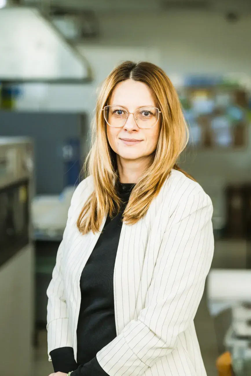 kobieta w białej marynarce, okularach, włosach blond stoi i założonymi dłońmi na tle laboratorium, PŁ, prof. Anna Masek