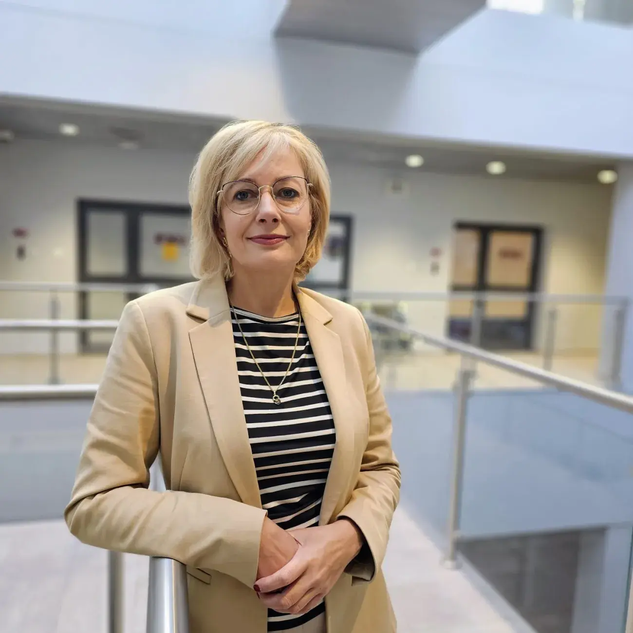 kobieta z blond włosami, w okularach, czerwonych ustach stroi oparta prawym łokcciem o barierkę nowoczesnego i jasnego wnętrzam prof. Hanna Kierzkowska-Pawlak