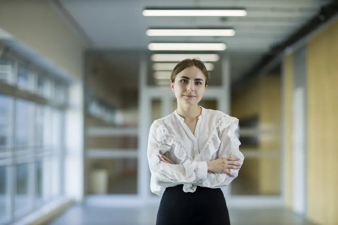 Izabela Kowalczyk, zdjęcie postaci stojącej z założonymi rękami, w białej ozdobnej bluzce, w tle korytarz