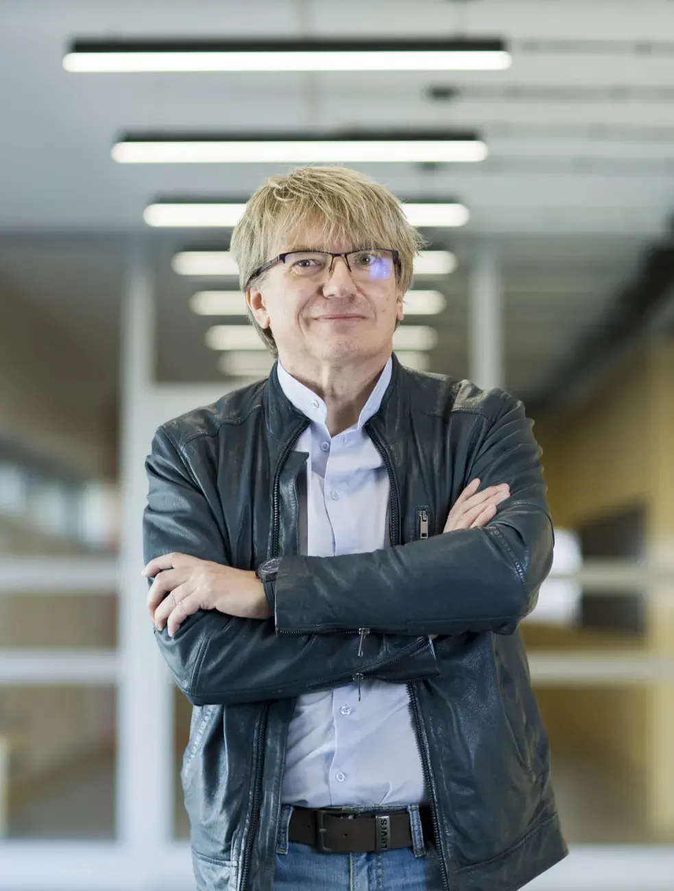 mężczyzna w średnim wieku, ubrany w skórzaną kurtkę i koszulę jasna, okulary, stoi z założonymi rękami na tle korytarza