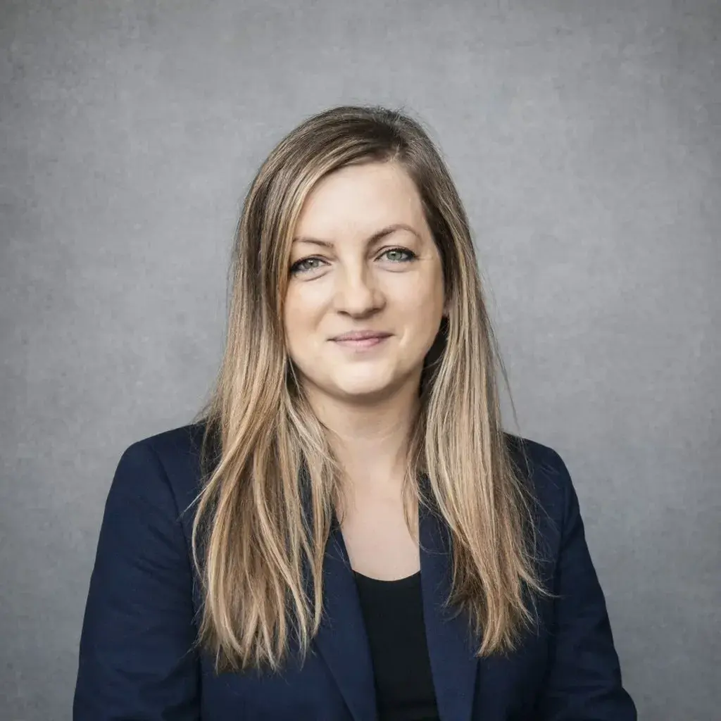 portret kobiety z długimi włosami, w ciemnym żakiecie, uśmiecha się, na tle szarej ściany, Joanna Oracz