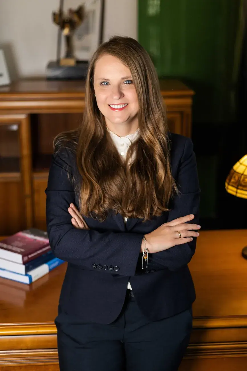prof. Marta Gmurek, elegancko ubrana, uśmiechnięta, z długimi włosami, opiera się z założonymi rękami o biurko