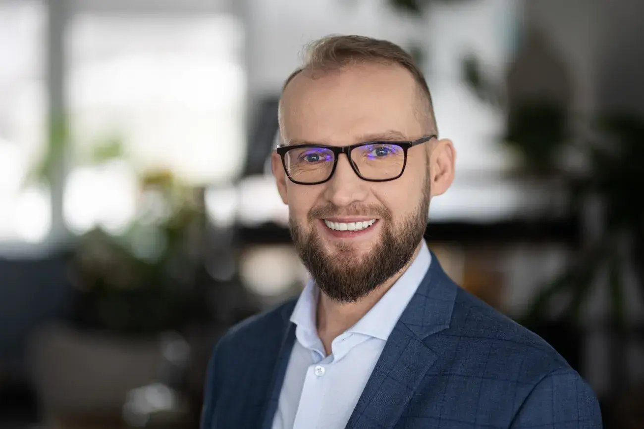 portret mężczyzny uśmiechniętego, w okularach, z brodą, w garniturze, w tle romyte biuro, Radosław Gajewski
