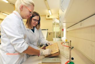 Zdjęcie portretowe: Dwie doktorantki z zespołu prof. Beaty Gutarowskiej ubrane w białe fartuchy stoją  w laboratorium przy blacie laboratoryjnym i dokonują pomiarów.