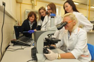Portrait photo: Prof. Beata Gutarowska's team of 5 in the laboratory. In the foreground a female scientist is looking through a microscope. In the background, three female PhD students and Prof. B. Gutarowska.
