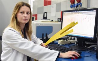 Portrait photo: Dr Eng. Anna Masek in a white coat is sitting at her desk in right profile. In her hand she is holding yellow stripes. On the right, a computer screen with data. In the background, a laboratory. 