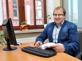 Portrait photo: Prof. Grzegorz Szymański in a blue jacket sits at his desk and computer.