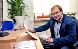 dr hab. inż. Grzegorz Szymański w niebieskiej marynarce siedzi przy biurku. W jednej ręce trzyma kartkę papieru, drugą ręką pisze na klawiaturze.