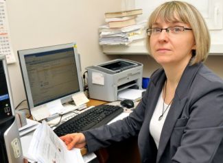 Portrait photo: Dr Eng. Janina Leks-Stępień is standing at a desk. Some documents, computer and printer in the background.