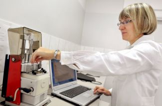 Portrait photo: Dr Eng. Janina Leks-Stępień in a white coat in left profile is standing at a specialist apparatus and a computer and is performing measurements.