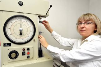 Zdjęcie portretowe: w białym fartuchu dr inż. Janina Leks-Stępień lewym profilem siedzi przy specjalistycznym urządzeniu.