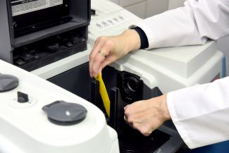 Overview photo. A fragment of a black-and-white device in a laboratory. In the foreground the hands of a female scientist in a white lab coat are inserting a yellow strip into a slot.