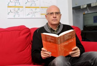 Portrait photo: Prof. Piotr Paneth is sitting on a red couch. In both hands he is holding an open book. A blackboard with chemical diagrams in the background.