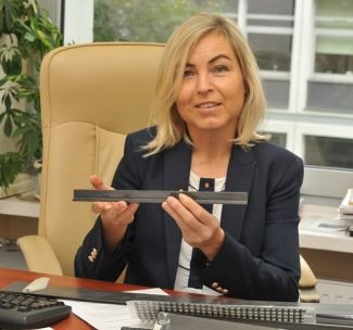 Portrait photo. Smiling Professor Renata Kotynia is sitting at her desk in her office. In her hands she holds a metal construction element.