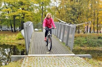 Prof. Renata Kotynia w stroju sportowym przejeżdża na rowerze przez most na kampusie Politechniki Łódzkiej. Dookoła zielone drzewa.